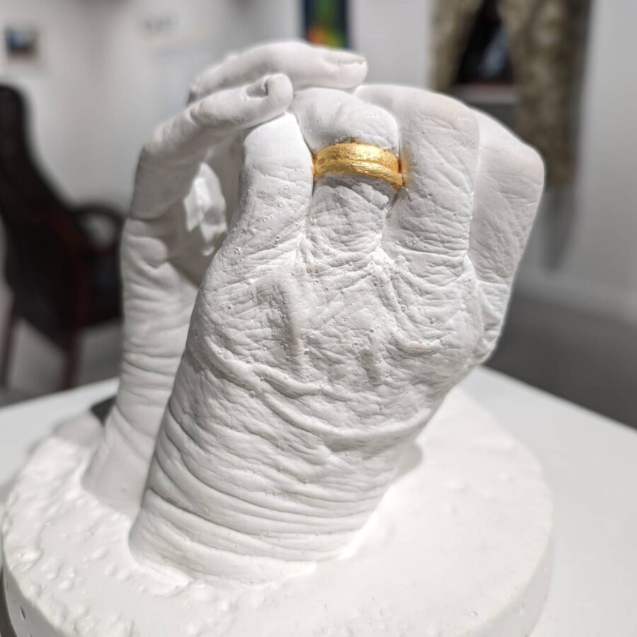 Plaster cast sculpture of an older woman's clasped hands with her wedding ring painted in gold.